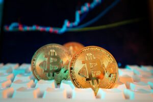 Close-up photo of hands clicking a mouse on a laptop keyboard with golden coins in the background symbolizing online investment in gold.
